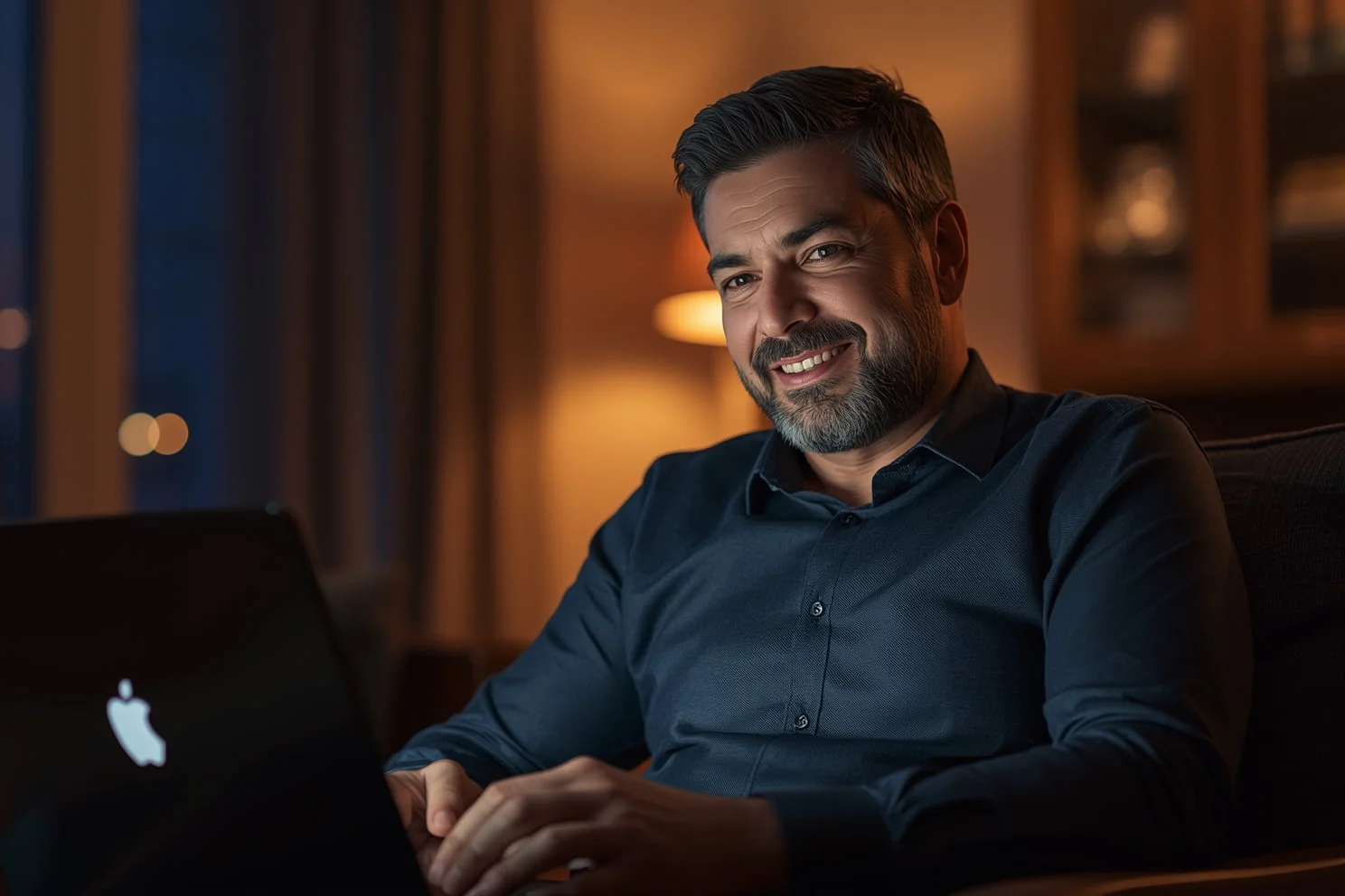 alt="Adult man sitting in an armchair in the evening, smiling while chatting online on a dating platform"