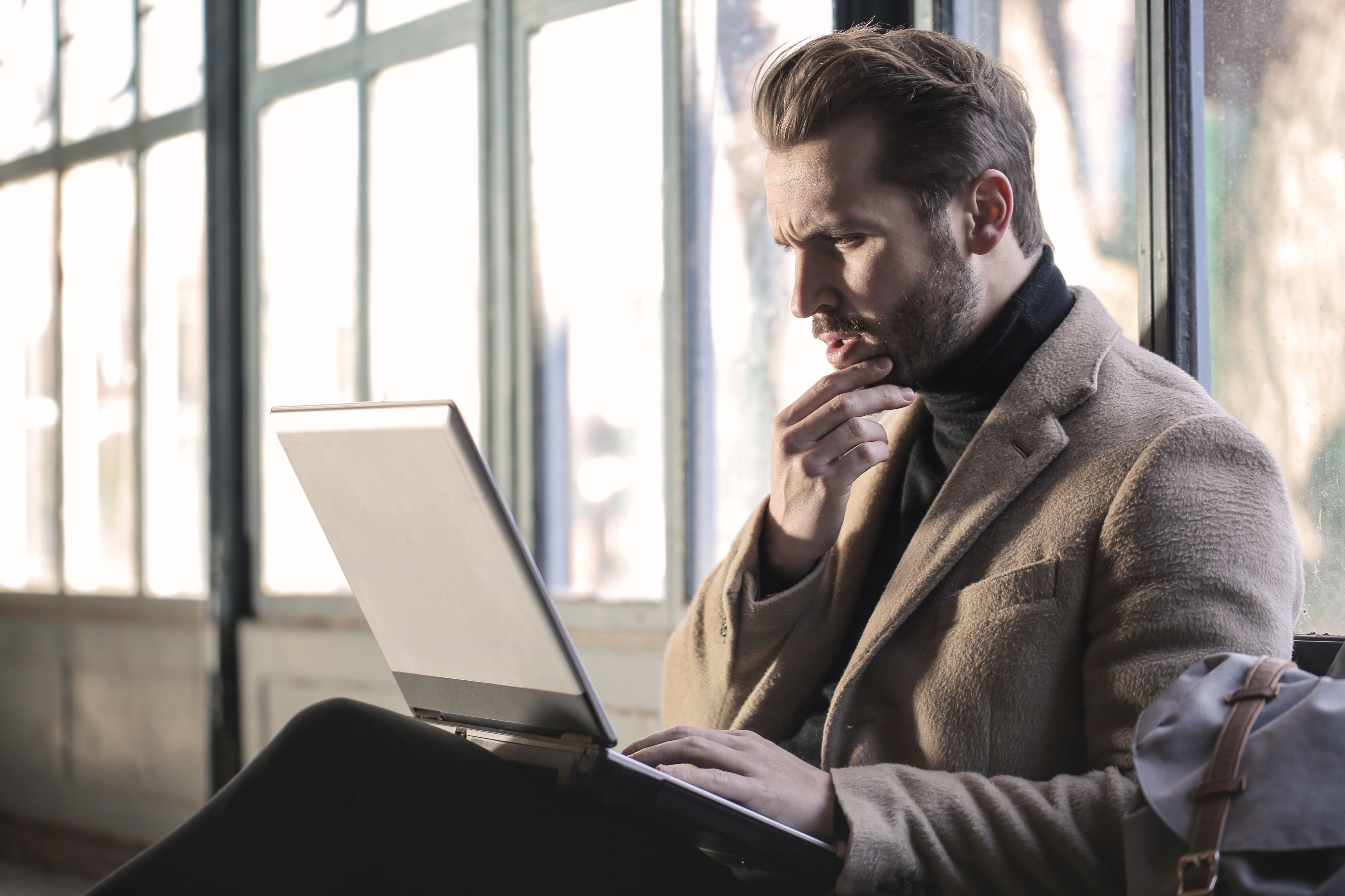 Man looking thoughtfully at a laptop while sitting by a large window.