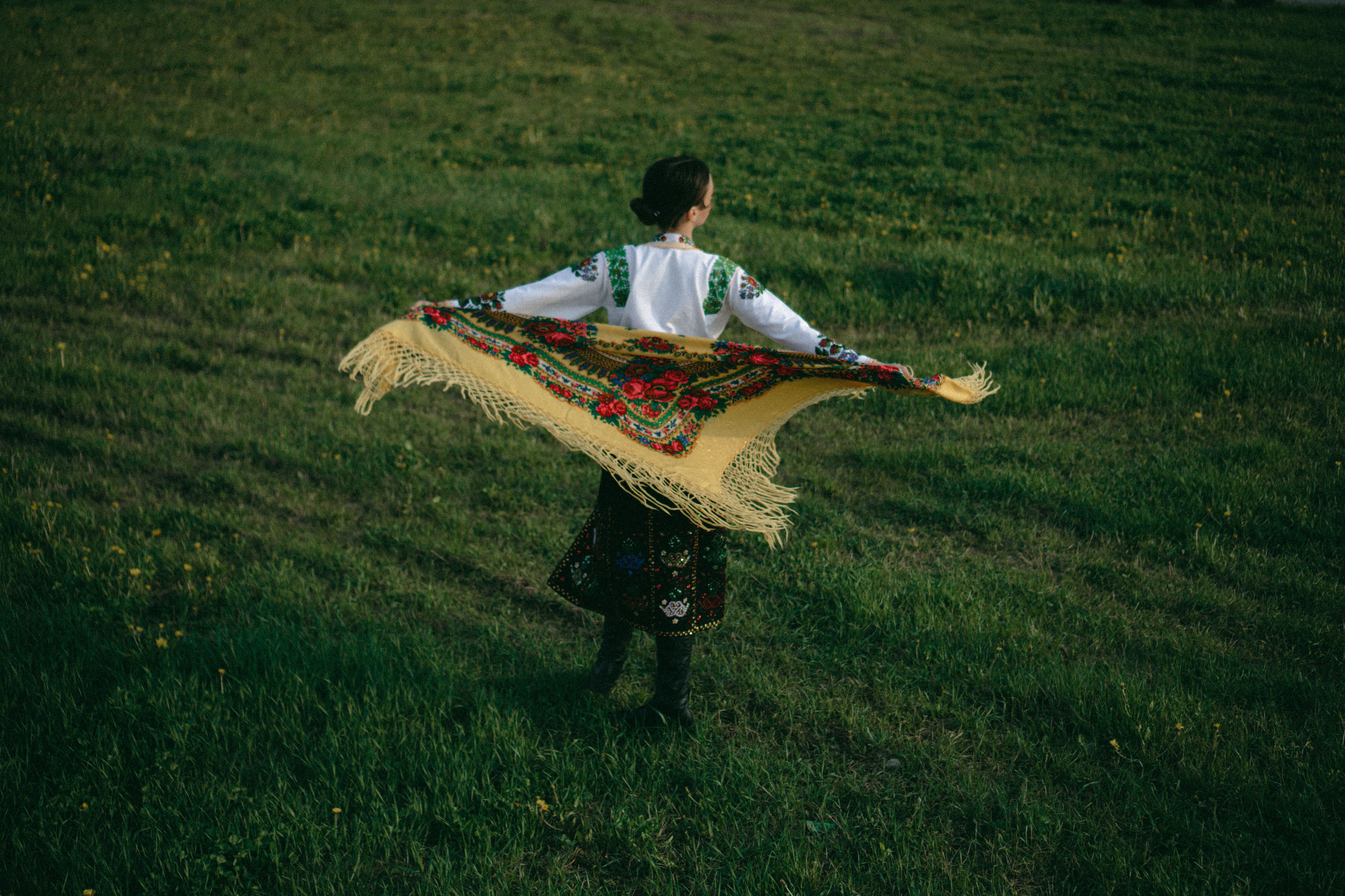 Back view of a woman in an embroidered shirt holding a bright floral shawl outdoors.